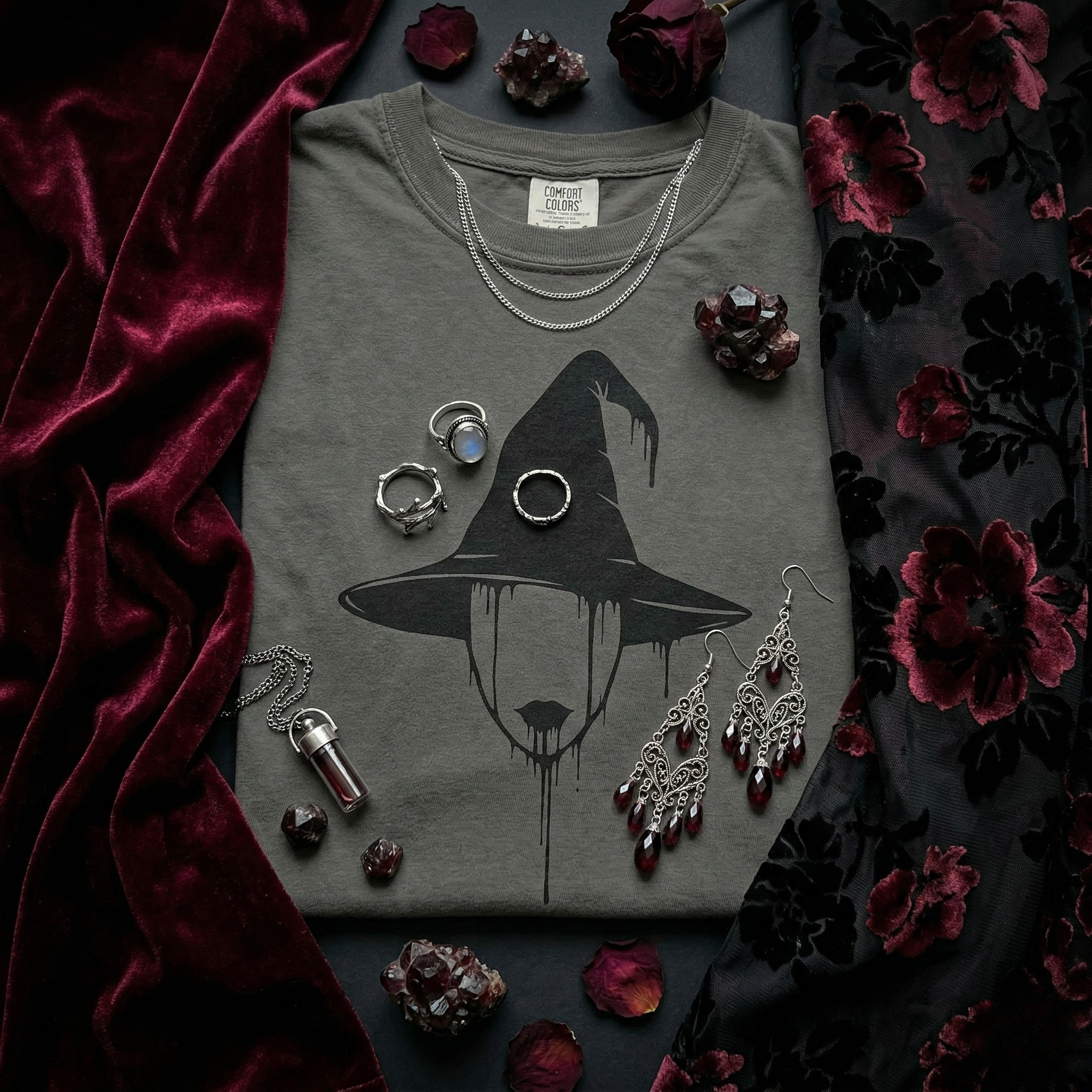 A close-up flat lay of a grey t-shirt on a black background, with a black graphic of a witch’s hat and a person with dripping black makeup on. The shirt is surrounded by rich textures: a burgundy velvet swatch and a dark floral fabric with black flowers. The composition features silver jewelry, including rings and earrings, and a small glass vial. The scene is illuminated by dark, romantic lighting with subtle shadows, and a color palette of wine, crimson, onyx, and silver.