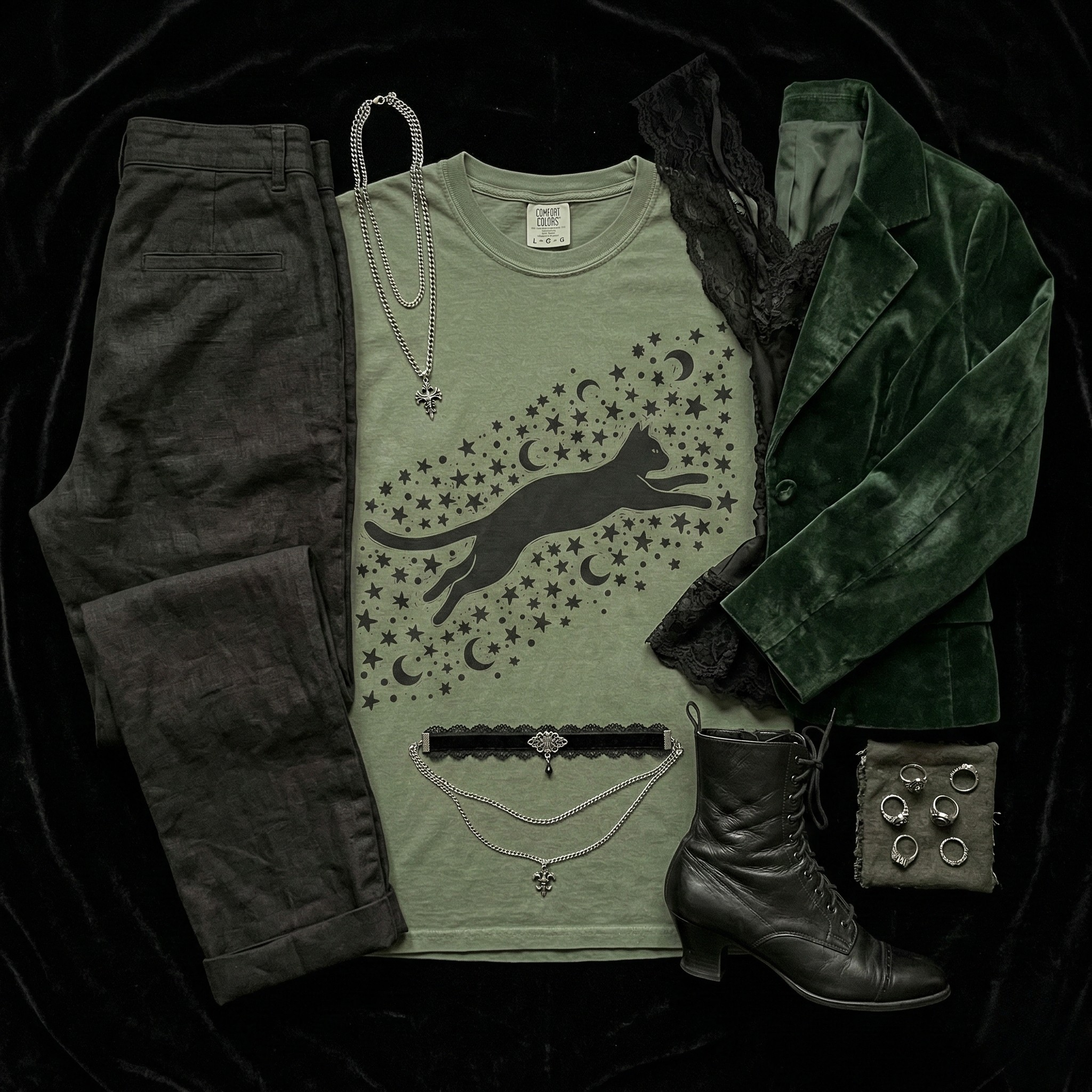 A curated flat lay outfit on a black velvet background. Center stage is an olive-green Comfort Colors t-shirt featuring a black graphic of a running cat within a constellation of stars and crescent moons. Surrounding the shirt are dark charcoal trousers on the left, and a dark green velvet blazer with a black lace detail on the right. Below these are a black leather Victorian lace-up boot, a collection of six silver rings, a layered silver choker necklace, and two tiered silver pendant necklaces. The entire arrangement has a dark romantic, capsule wardrobe feel.